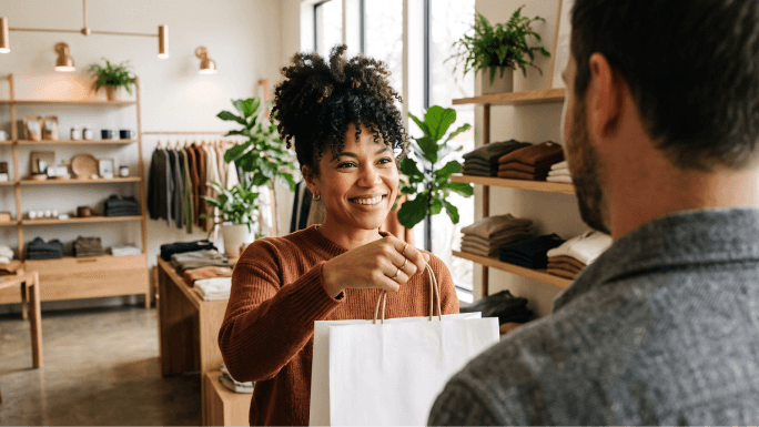 Small business owner handing a boutique purchase bag to a shopper at checkout.