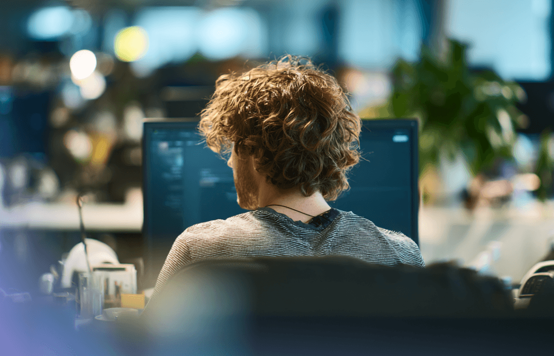 Over-the-shoulder view of a developer with curly hair working on a computer in a modern office.